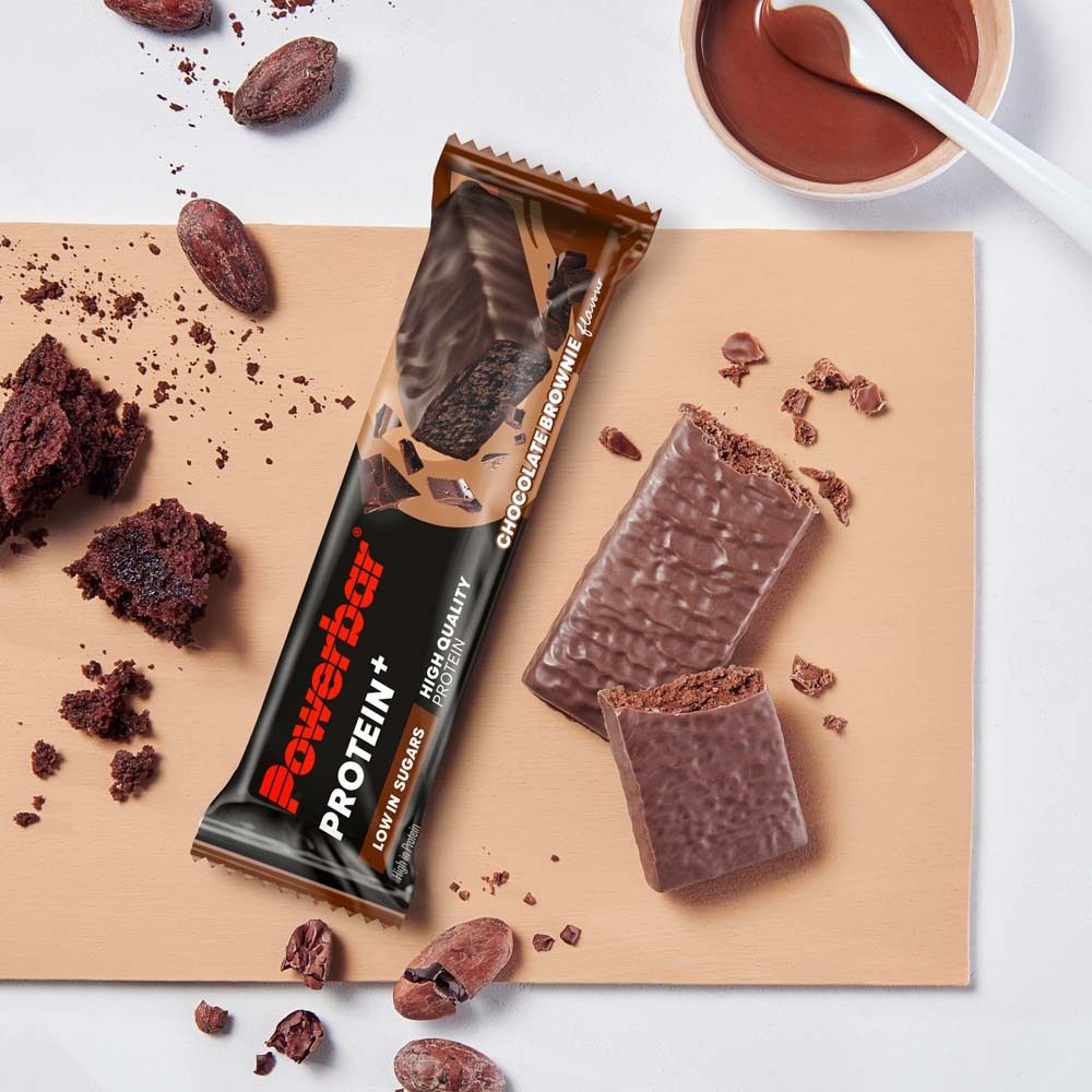 Powerbar PROTEIN+ LOW SUGAR CHOCOLATE BROWNIE. Barre et morceaux sur planche de bois. Fèves de cacao et sauce au chocolat.
