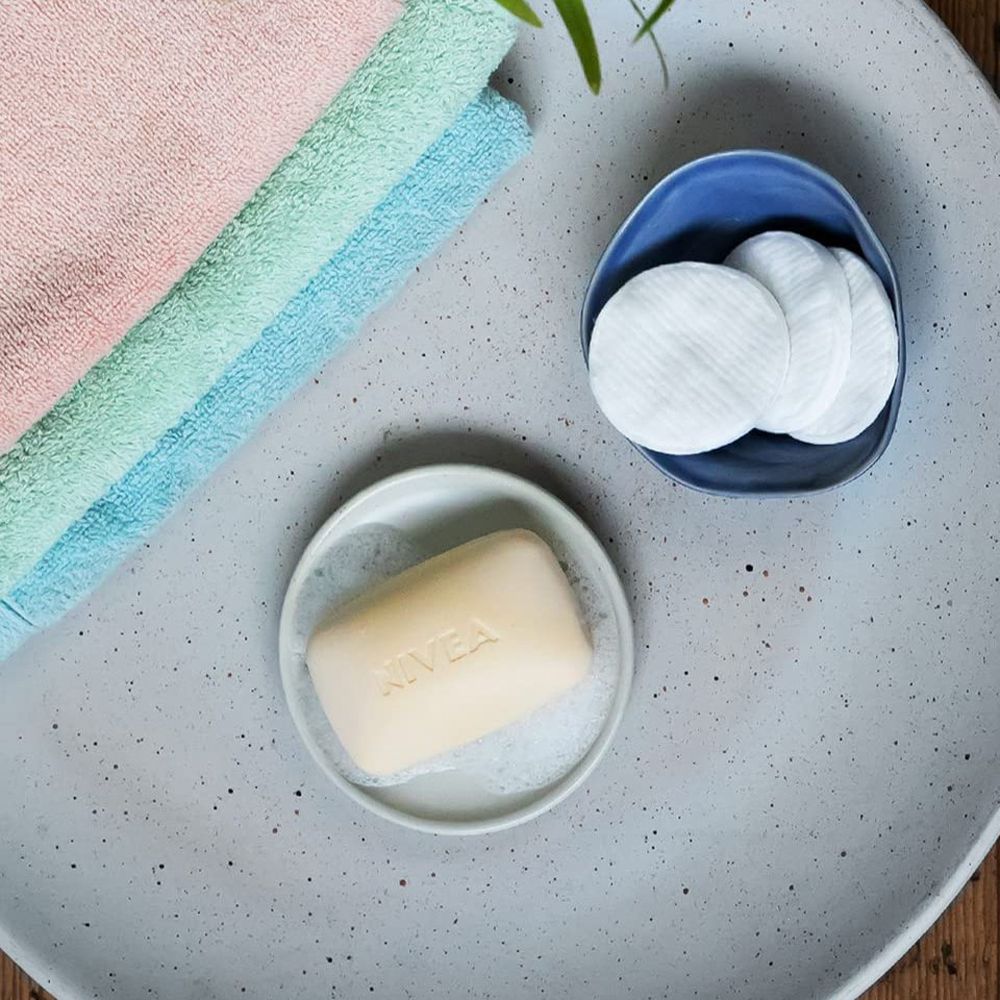 Savon NIVEA Creme Soft sur un porte-savon, entouré de mousse. À côté se trouvent des cotons démaquillants et des serviettes.