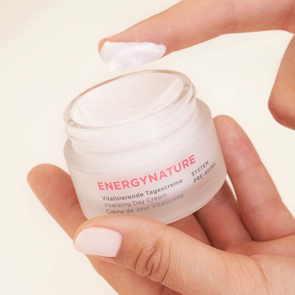 Main tenant un pot de crème. Inscription ENERGYNATURE, Crème de Jour Vitalisante. Un doigt touche la crème.