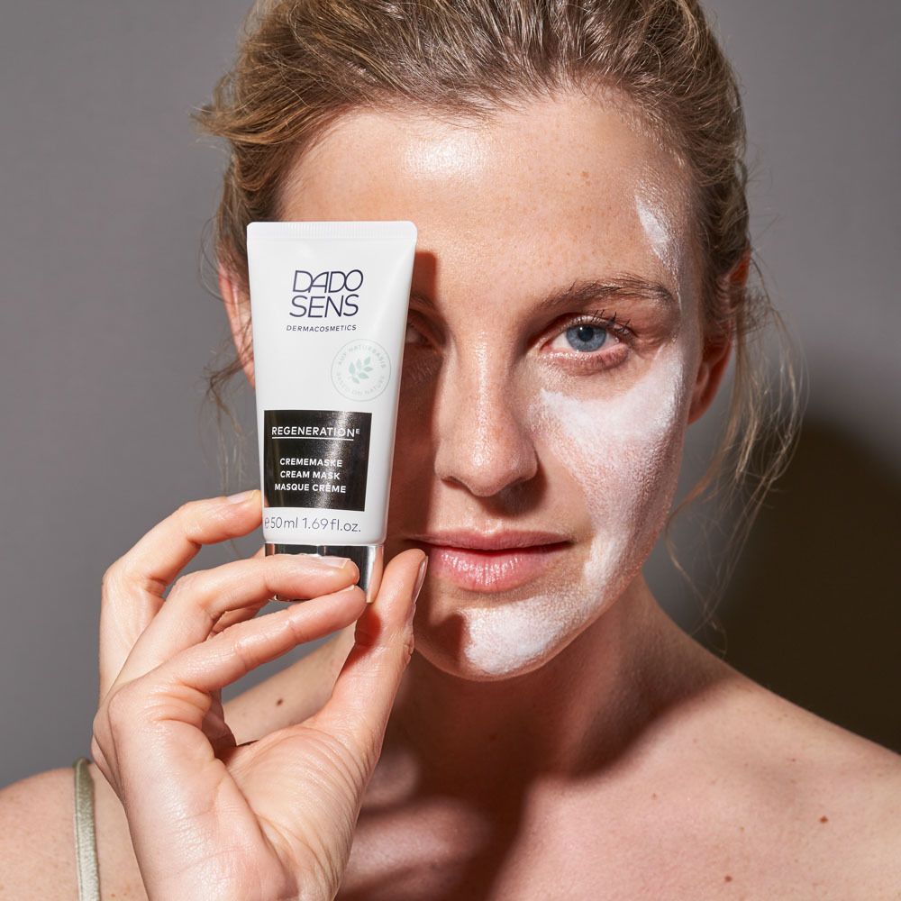 Femme avec crème blanche sur le visage. Tient un tube DADO SENS REGENERATION. Fond gris.