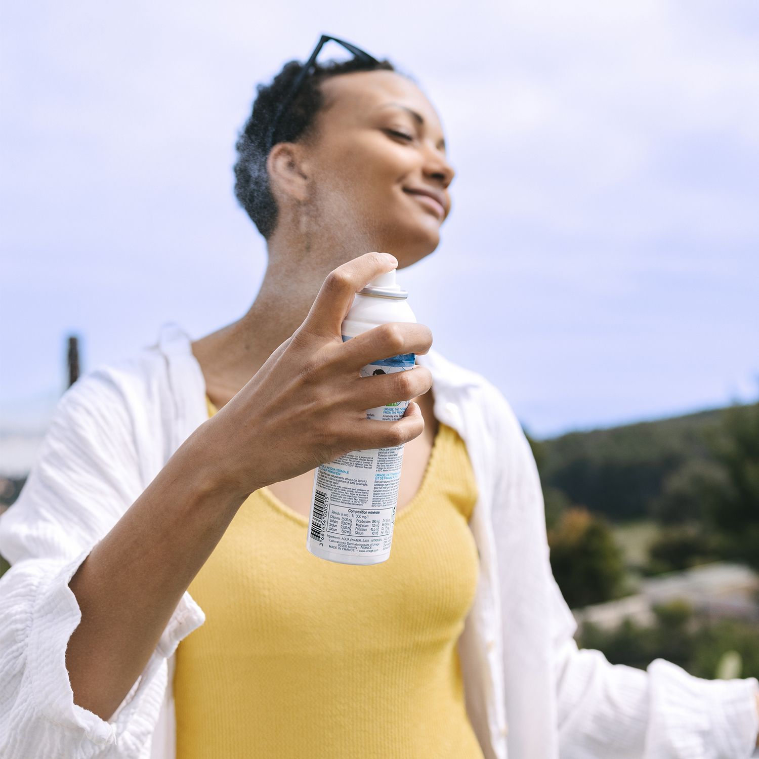 Femme vaporisant Uriage Eau Thermale sur son visage. Flacon blanc, bouchon bleu. Arrière-plan : nature.