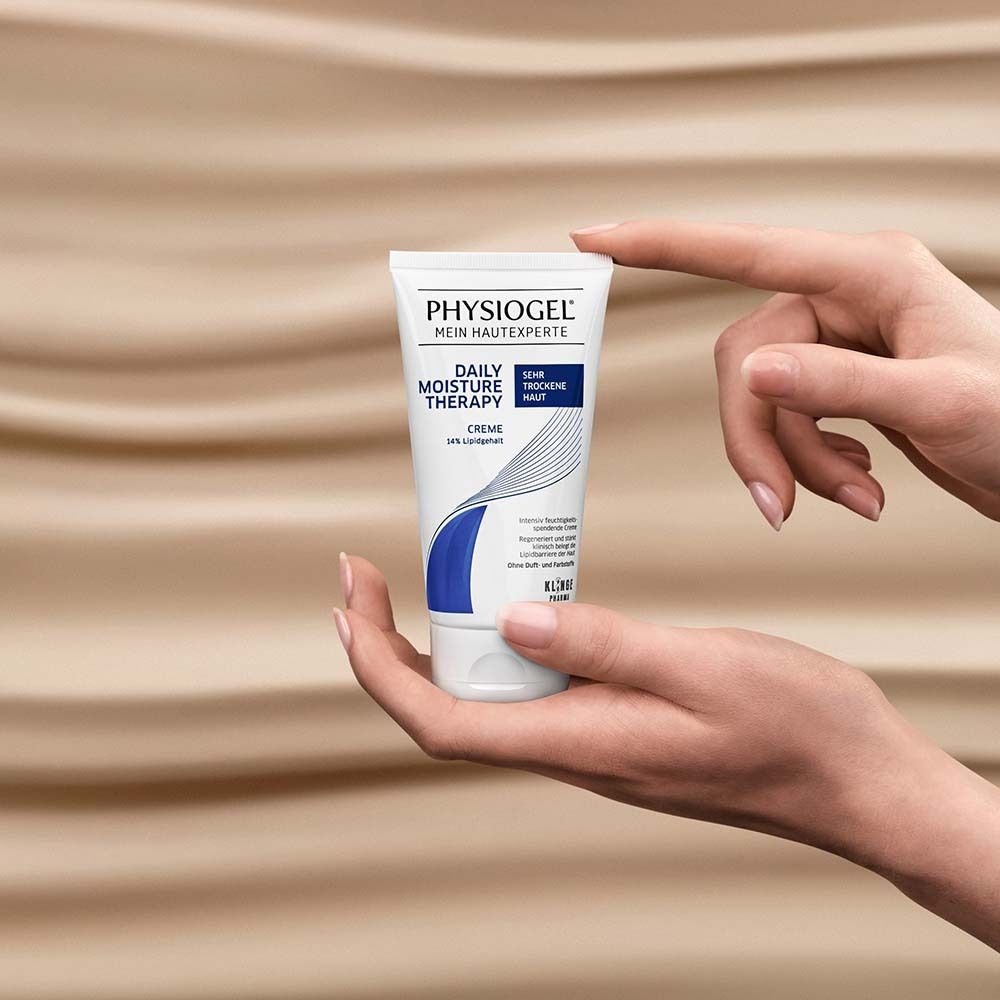 Mains tenant un tube de crème PHYSIOGEL® Daily Moisture Therapy. Blanc, accents bleus. Fond brun clair.