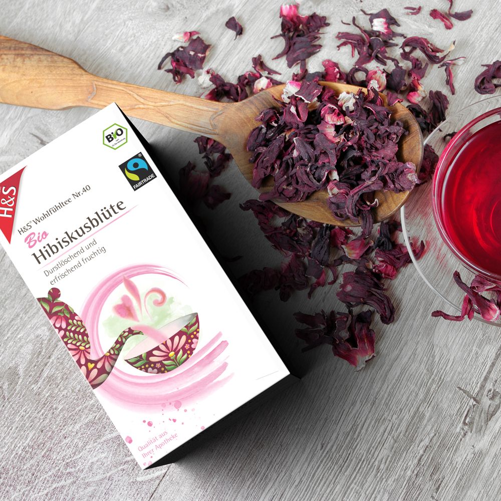 Boîte de thé H&S Bio à l'hibiscus à côté d'une cuillère en bois avec fleurs séchées et une tasse de thé.