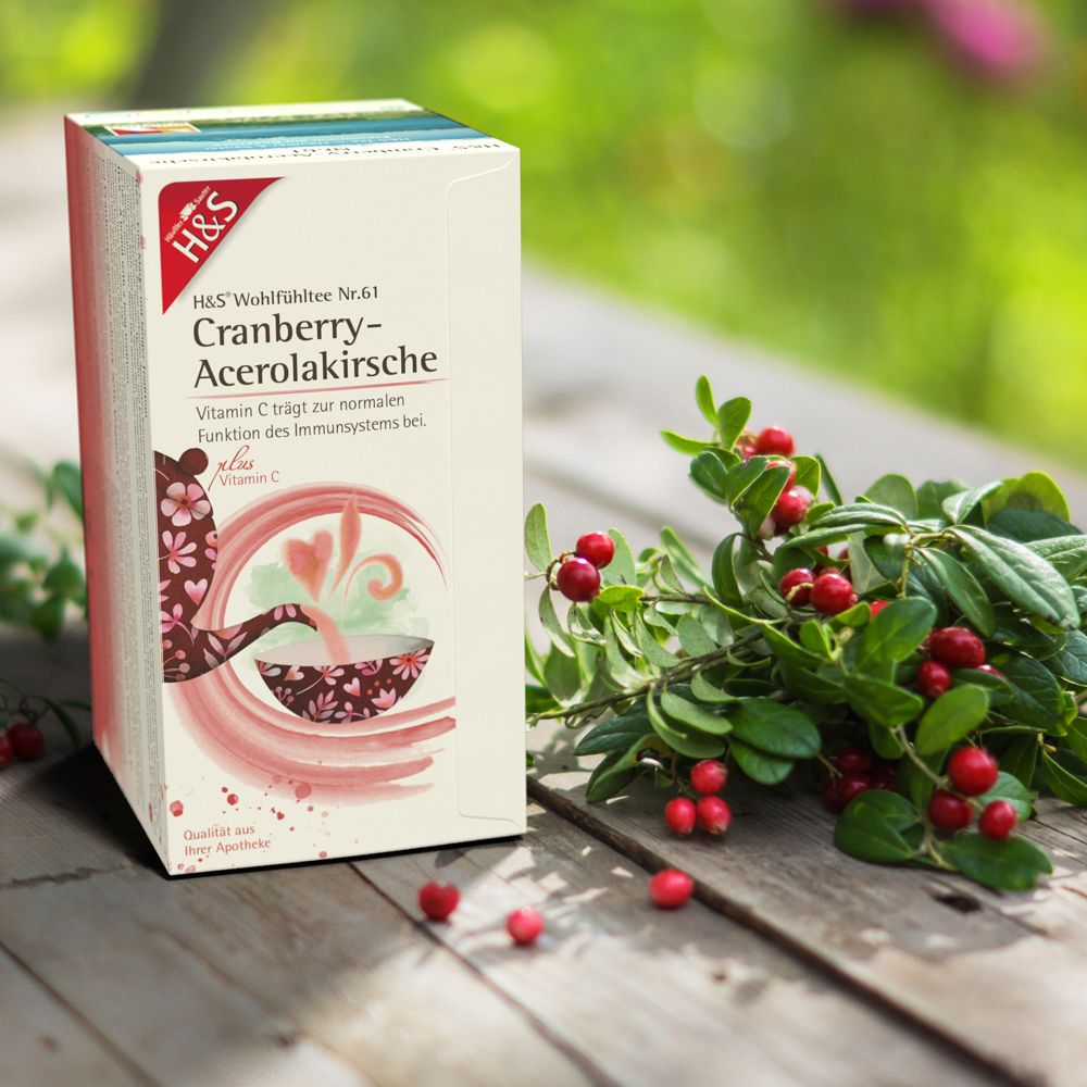 Boîte H&S Cranberry-Acerolakirsche Wohlfühltee à côté de canneberges fraîches sur surface en bois.