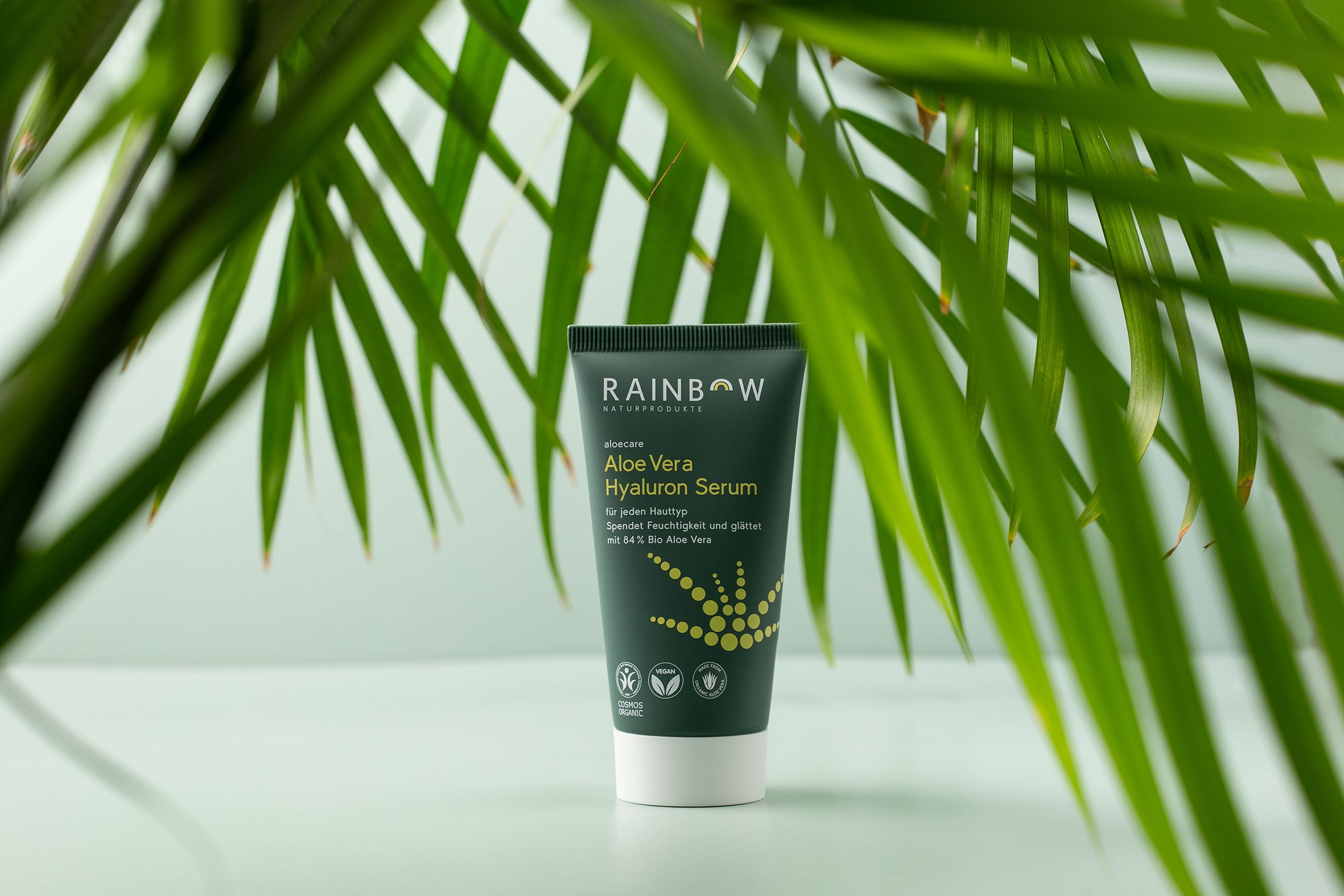 Tube vert devant des feuilles. Produit: Aloe Vera Hyaluron Serum. Labels vegan et bio. Fond vert.