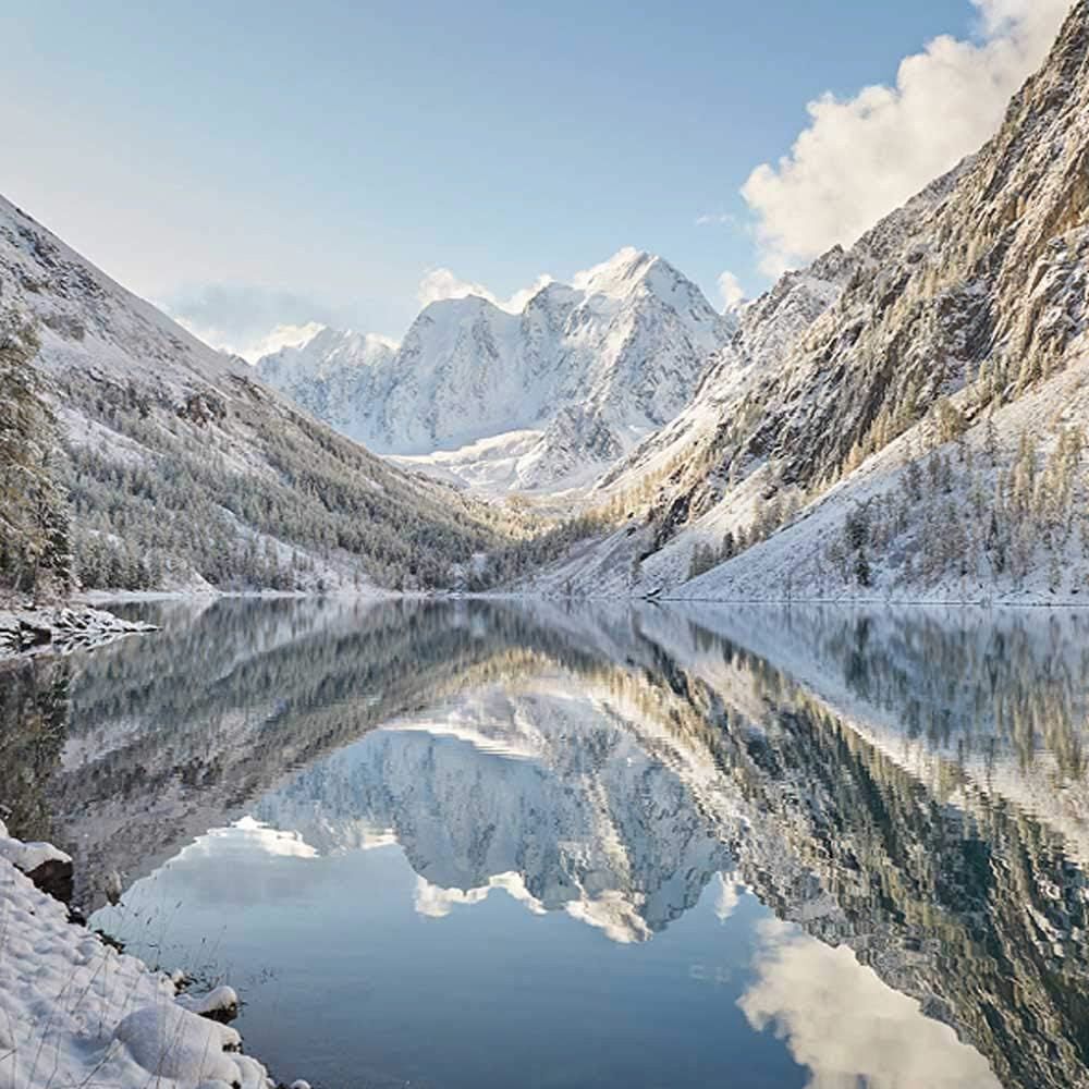 Surface de lac de montagne lisse comme un miroir reflétant des montagnes enneigées et des arbres sous un ciel bleu avec des nuages.