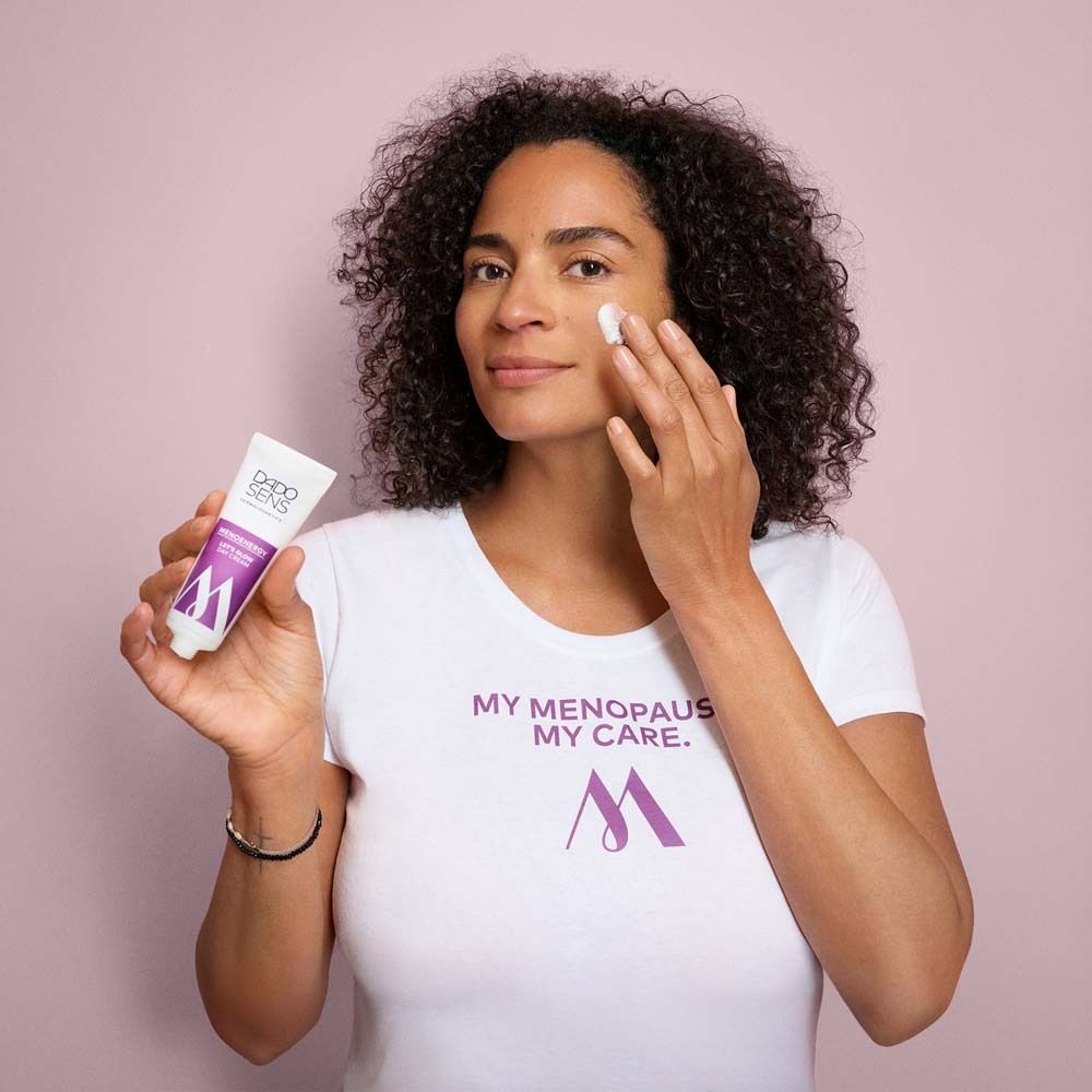 Femme aux cheveux bouclés tenant un tube. T-shirt blanc avec logo. Crème sur le visage. Fond rose.