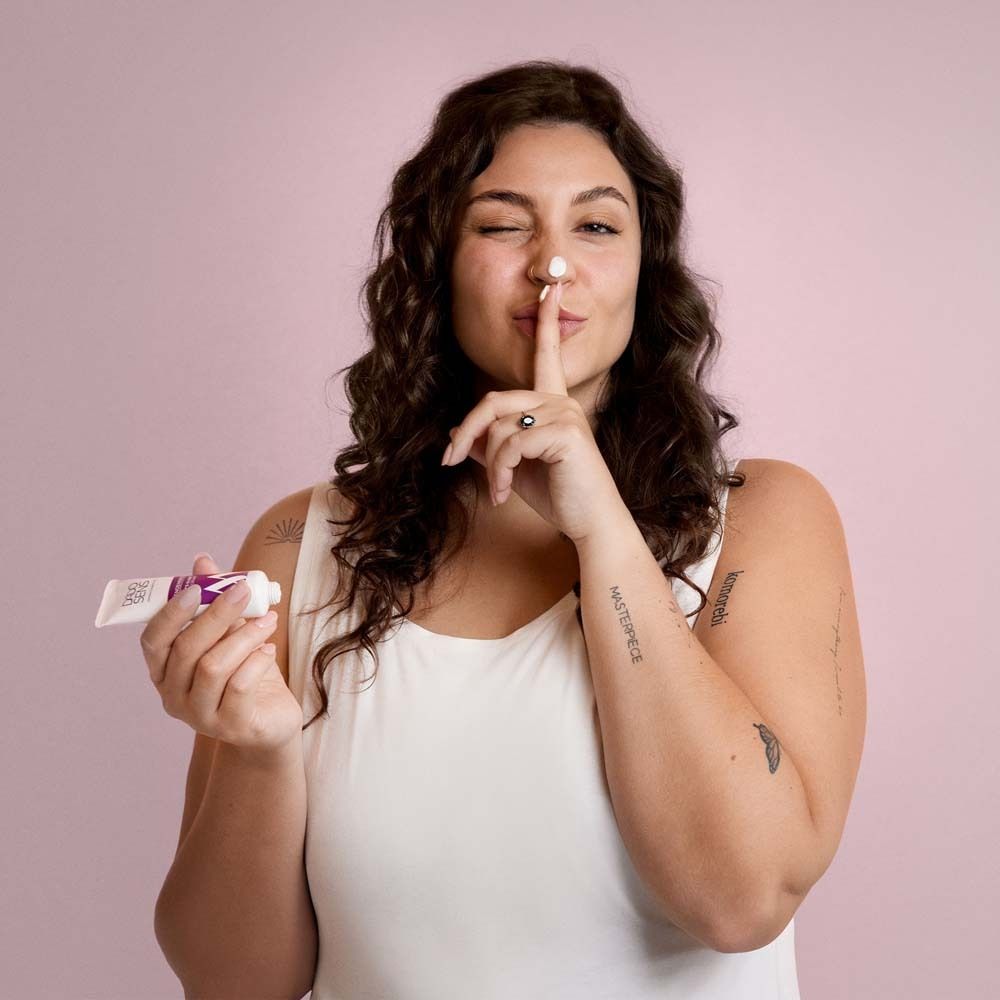 Femme aux cheveux bouclés tenant un tube. Doigt devant la bouche. Crème sur le nez. Fond rose.