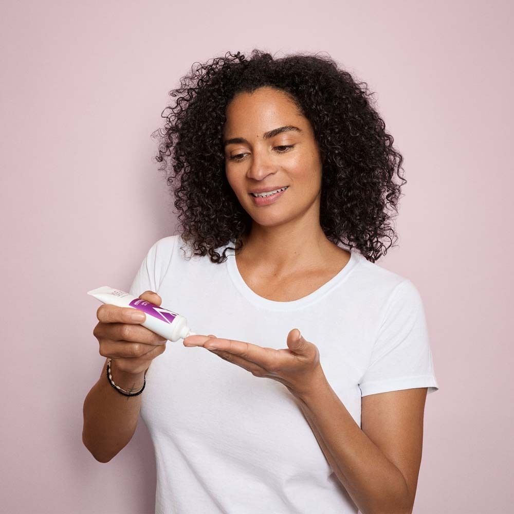 Femme aux cheveux bouclés tenant un tube. Elle dépose du produit sur sa main. Fond rose.