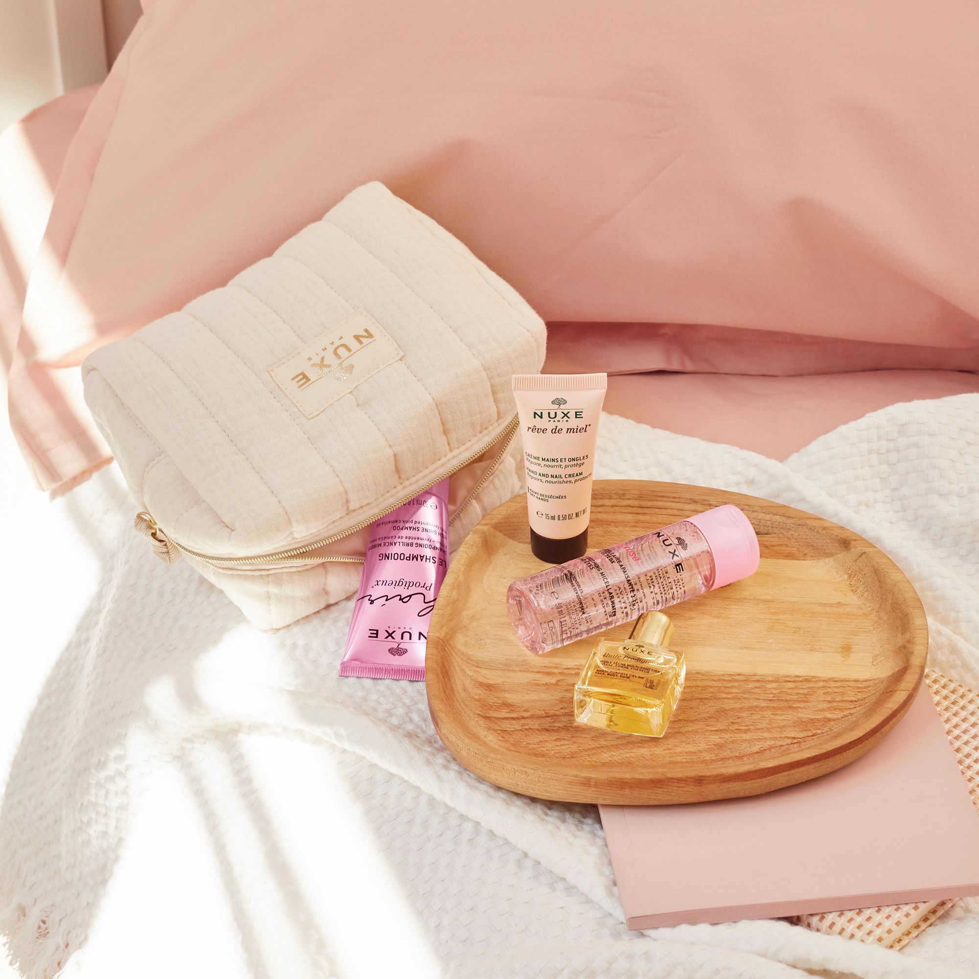 Trousse de toilette et produits sur un plateau en bois. Contient shampooing, huile, spray visage et crème mains. Sur linge de lit rose.
