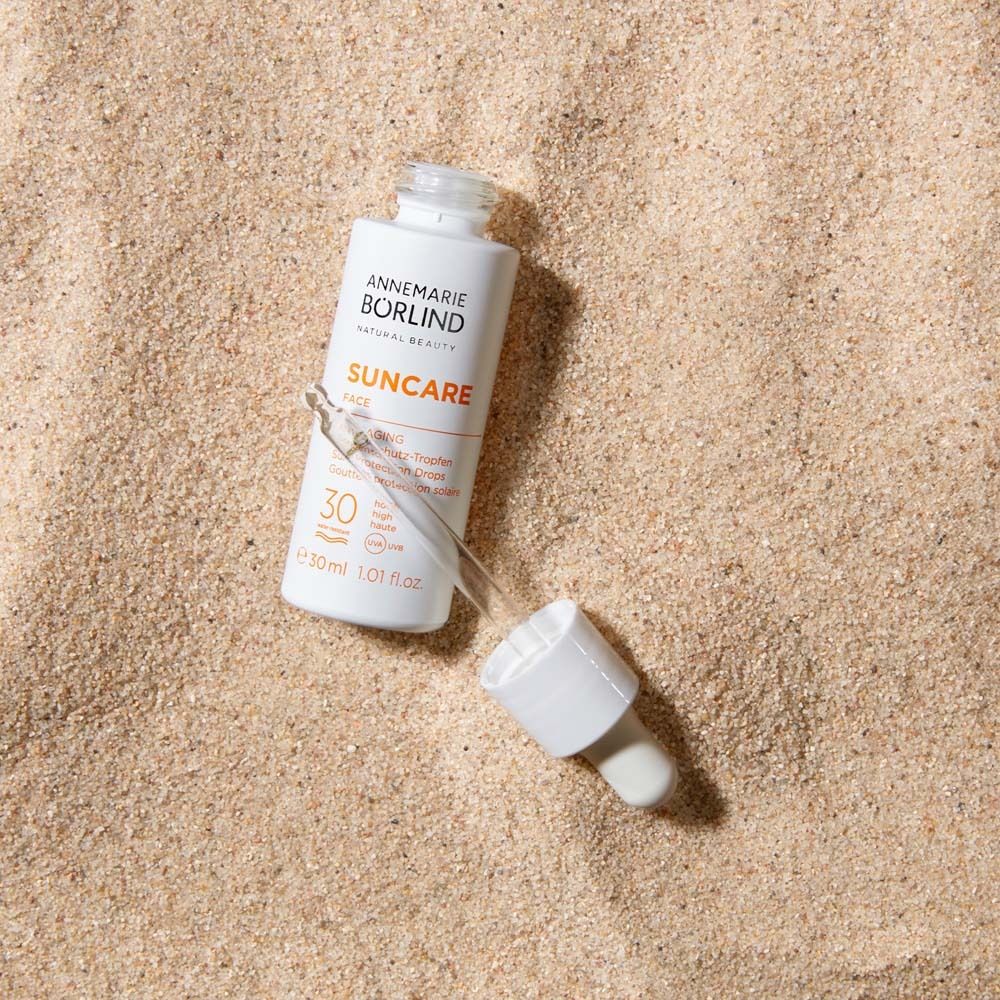 Flacon blanc avec pipette sur sable. Inscription : Suncare Face, Anti-Âge, SPF 30.