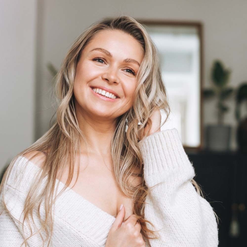 Portrait d'une femme blonde souriante avec de longs cheveux. Elle porte un pull blanc. L'arrière-plan est flou.