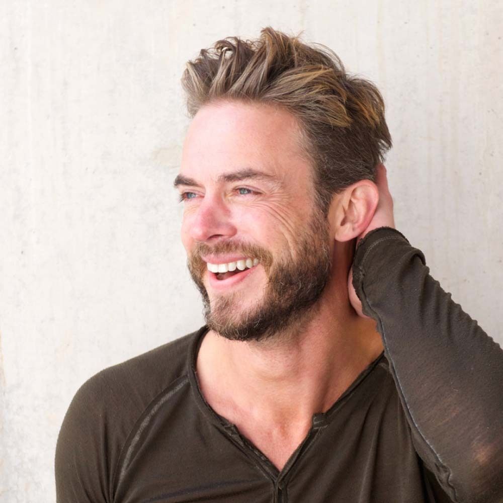 Un homme souriant avec une barbe et des cheveux blonds. Il touche l'arrière de sa tête. Le fond est gris clair.