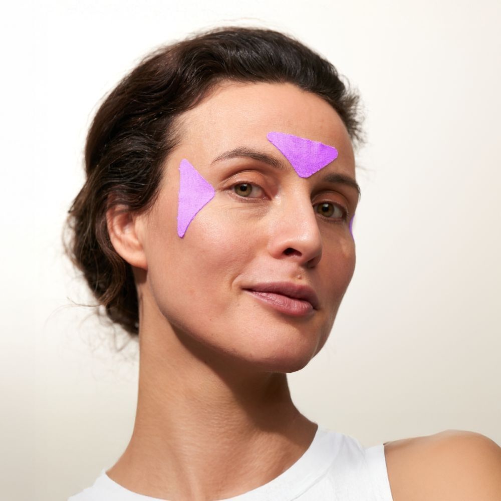 Femme avec deux patchs faciaux violets sur le front. Nom du produit non visible.