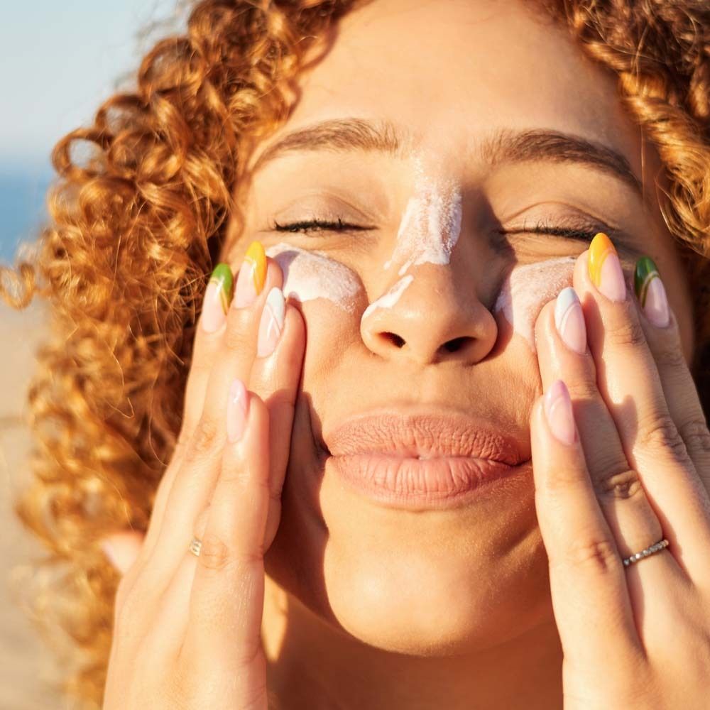 Femme appliquant de la crème solaire sur son visage. Peau claire, cheveux bouclés, ongles colorés. Gros plan.