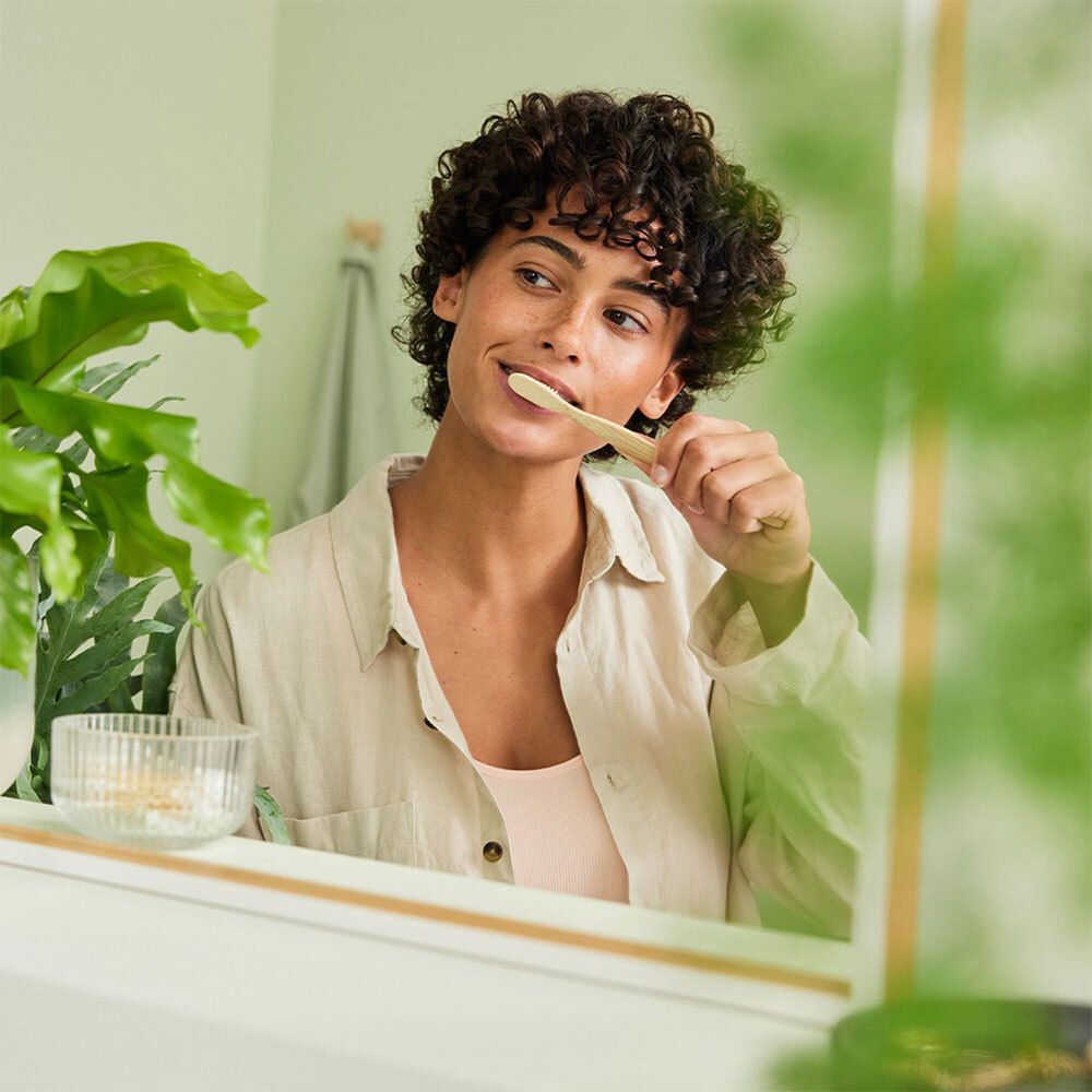 Femme se brosse les dents devant un miroir. En arrière-plan, pot de dentifrice. Brosse à dents dans la bouche.