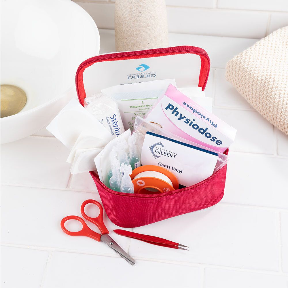 Trousse de secours rouge ouverte avec contenu. Ciseaux, pince à épiler, pansements, gants, sparadrap, sérum. Fond: lavabo.