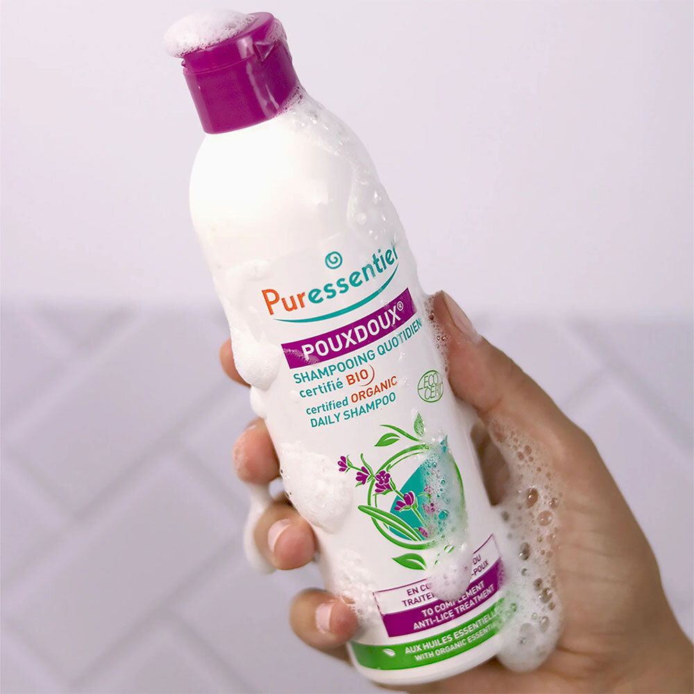 Flacon de shampoing blanc, bouchon violet, tenu par une main. Mousse recouvrant le flacon. Inscription : POUXDOUX.