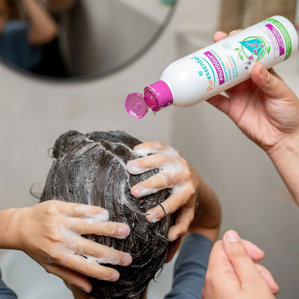 Personne se lave les cheveux avec du shampoing. Flacon avec bouchon violet tenu. Inscription : POUXDOUX.