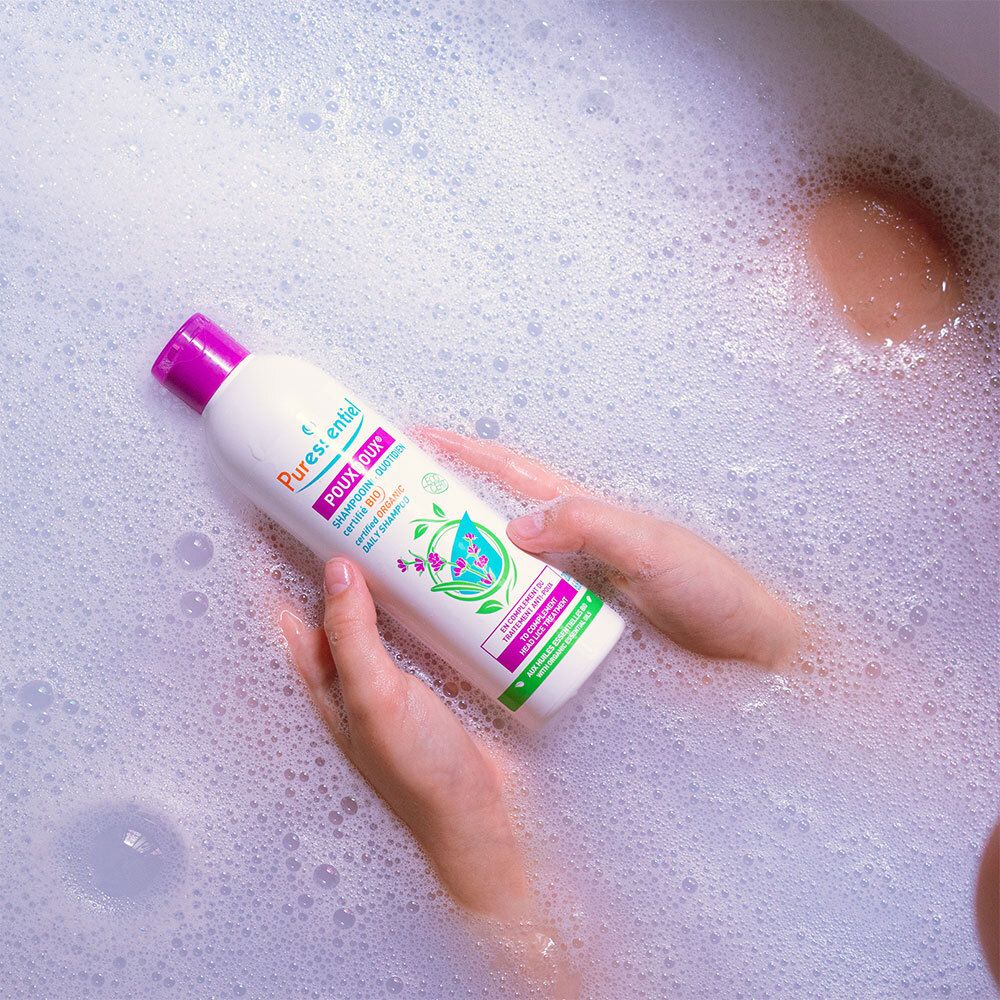 Personne dans une baignoire, flacon de shampoing dans les mains. Mousse. Flacon avec bouchon violet. Inscription : POUXDOUX.