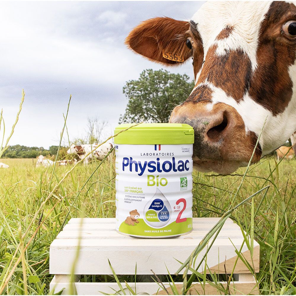 Boîte Physiolac Bio 2 sur planche en bois dans l'herbe. Vache en arrière-plan. Certification AB. 6/12 mois.