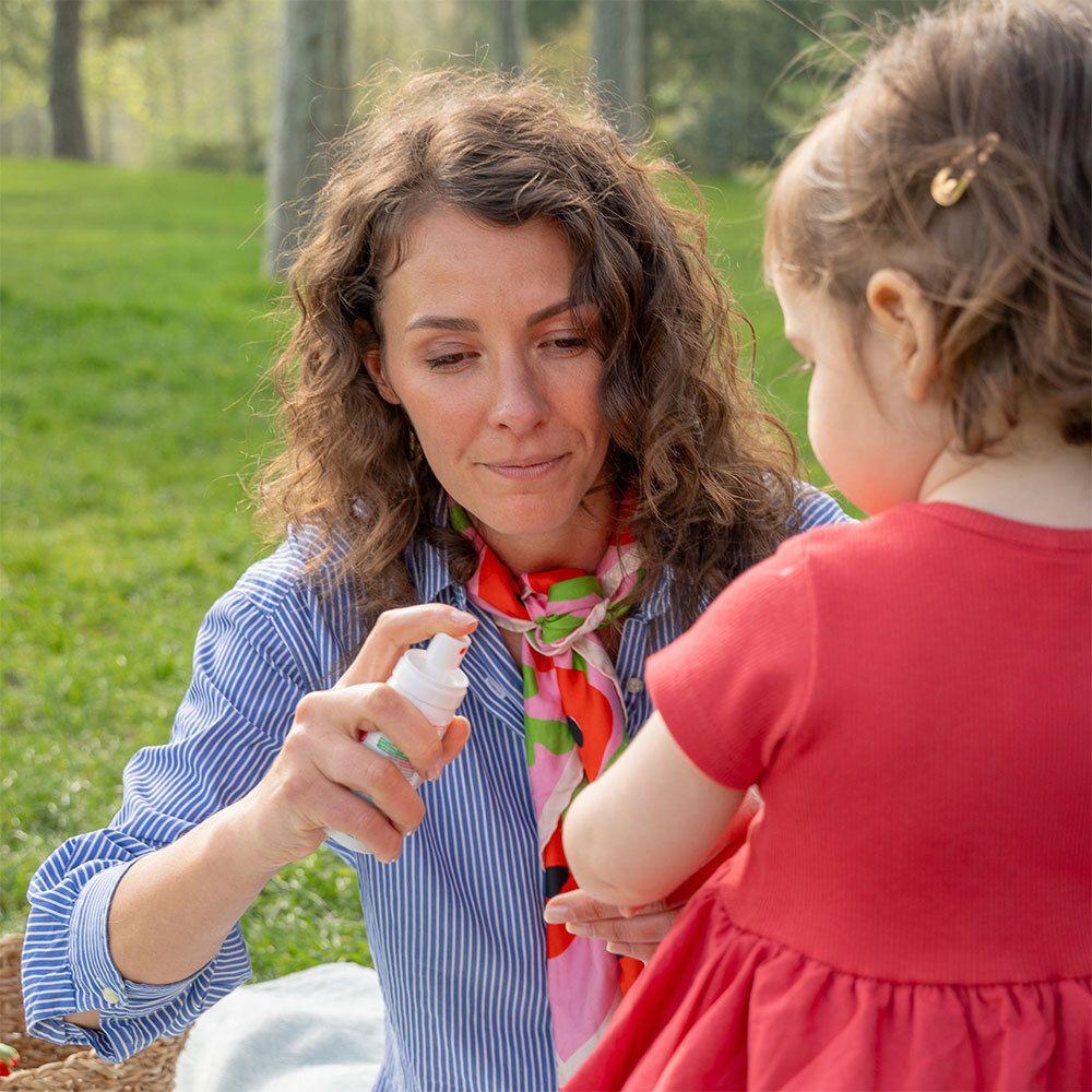 Femme vaporisant sur le bras d'un enfant. Spray rouge et blanc. Inscription: Anti-Pique Spray Répulsif Bébé.