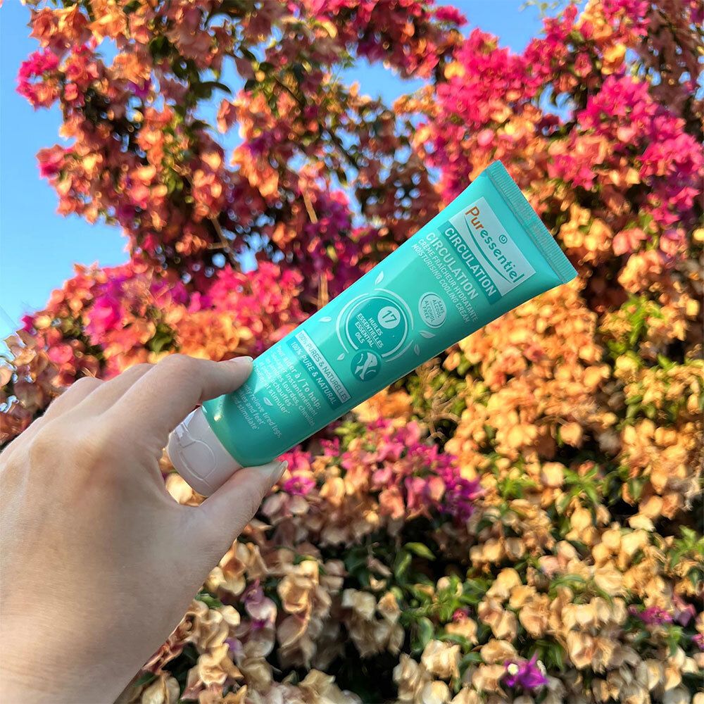 Main tenant un tube turquoise devant des fleurs. Inscription : Puressentiel Circulation. Contient 17 huiles essentielles. Sans alcool.