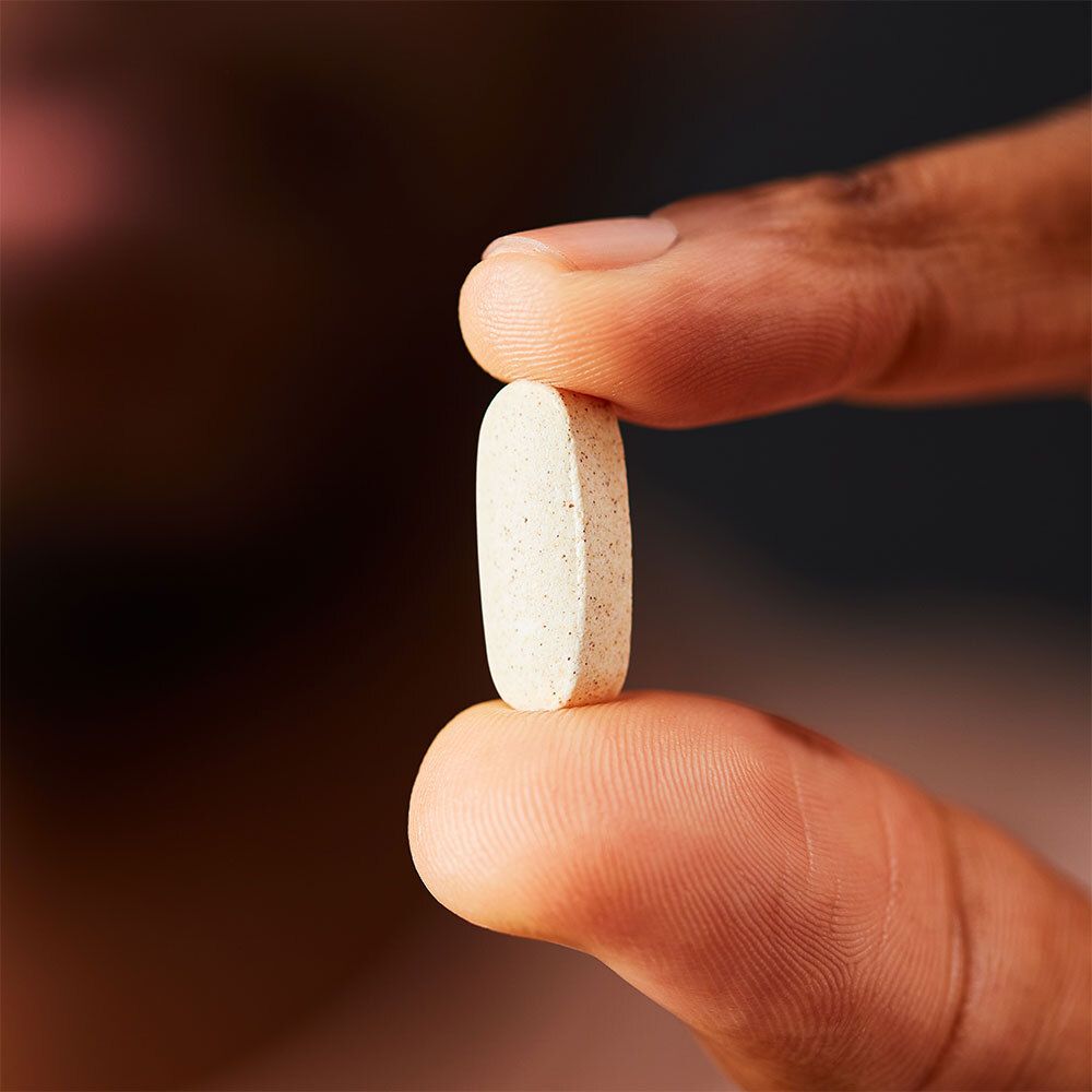 Comprimé beige, oblong, tenu entre les doigts. Bords arrondis.