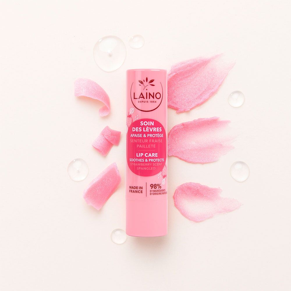 Stick de soin des lèvres rose avec éclaboussures roses et gouttes d'eau. Logo LAINO, texte: SOIN DES LÈVRES, APAISE & PROTÈGE, SENTEUR FRAISE, PAILLETÉ.