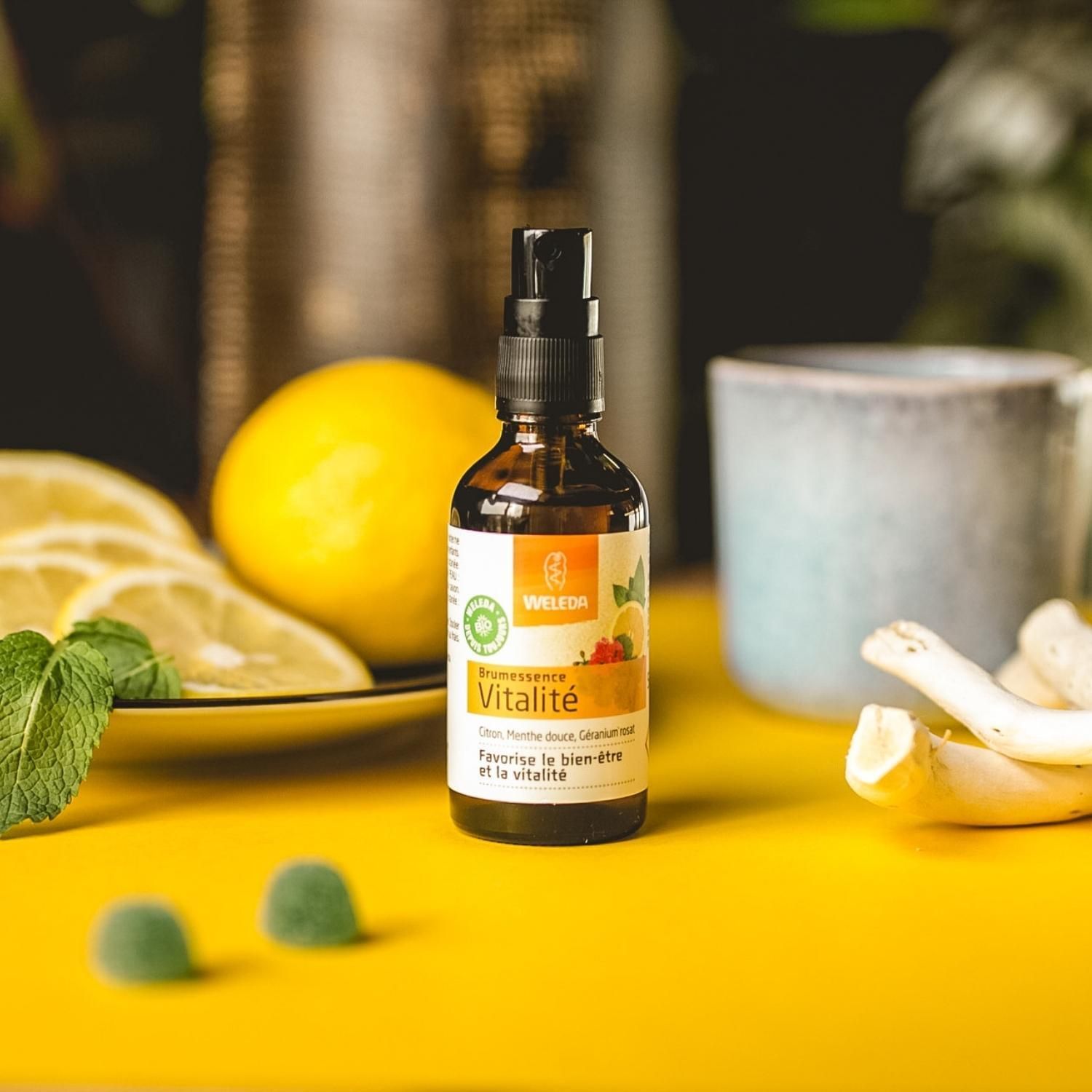 WELEDA Brumessence® Vitalité. Flacon sur fond jaune. Citron, menthe, géranium. Avec citron, tasse, feuilles.