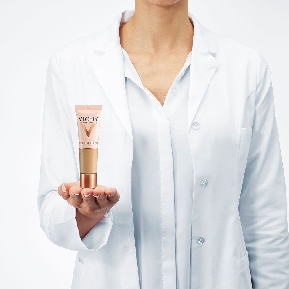 Personne en blouse blanche tenant un tube de fond de teint. Logo Vichy et Mineralblend.