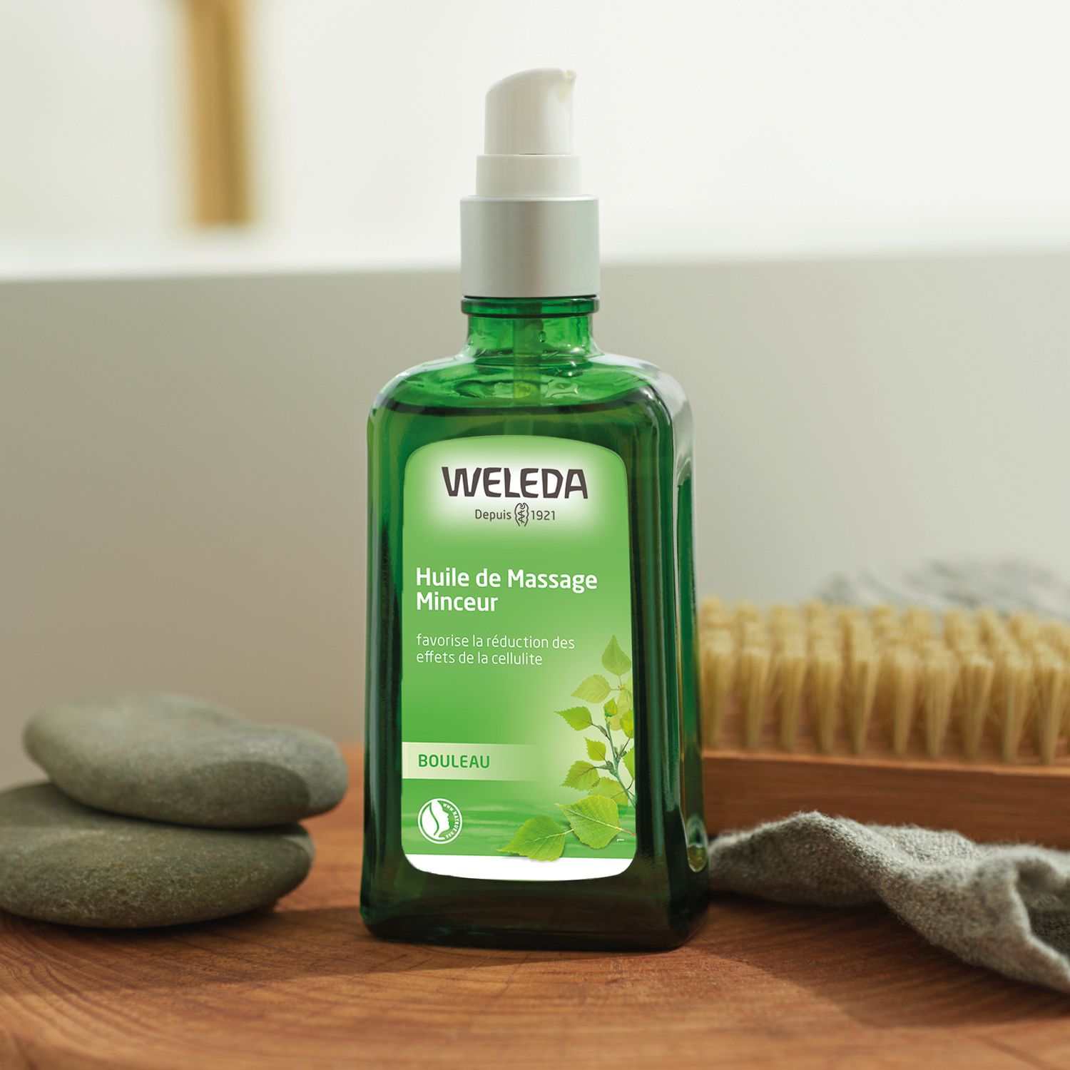 Flacon vert avec vaporisateur. Inscription: WELEDA, Huile de Massage Minceur, BOULEAU. Pierres et brosse à côté.