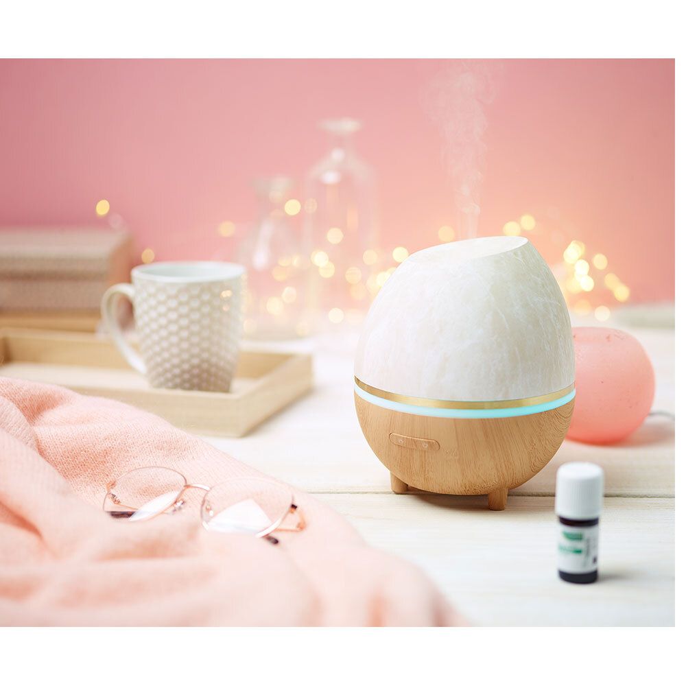 Diffuseur ovoïde sur une table. Base en bois, partie supérieure blanche. Accessoires : tasse, lunettes, flacon d'huile.