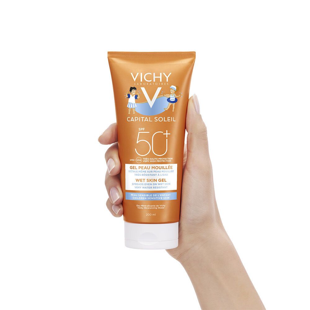 Main tenant un tube de crème solaire orange avec bouchon blanc. Inscriptions : VICHY, Capital Soleil, SPF 50+.