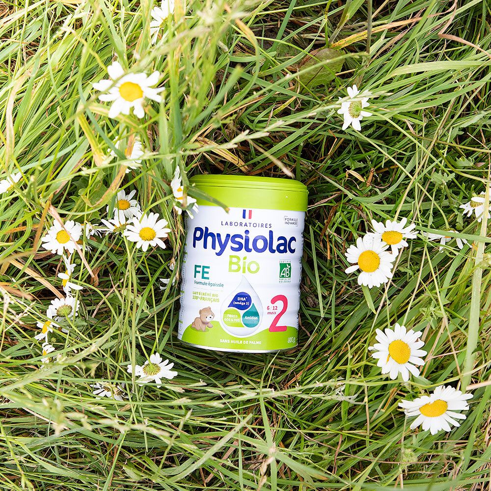 Boîte Physiolac Bio 2 dans l'herbe et les marguerites. Couvercle vert, contenant blanc. Inscriptions : Bio, 2, 6/12 mois.