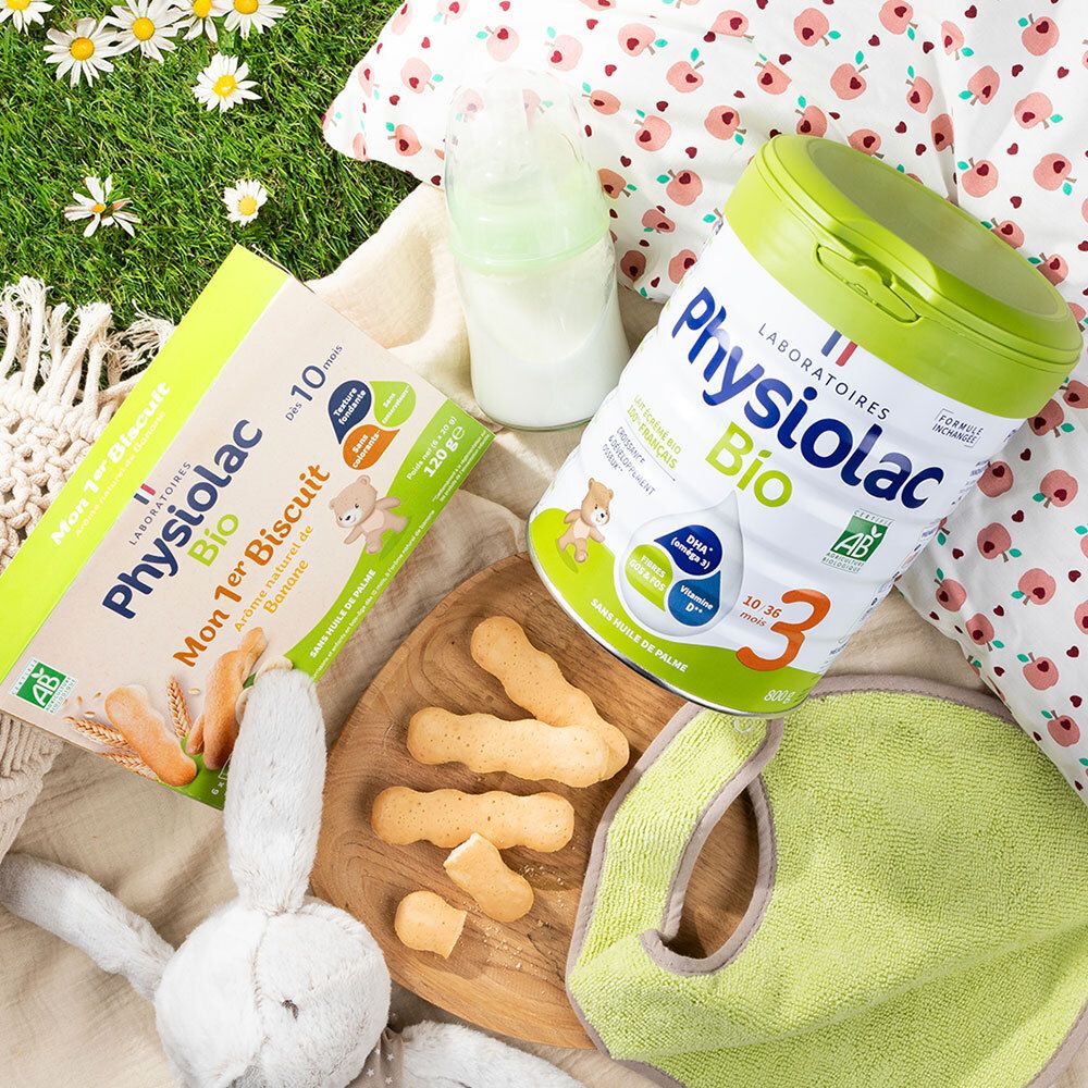 Scène avec produits Physiolac Bio. Mon 1er Biscuit Banane, lait en poudre, biberon, bavoir et jouet sur l'herbe.