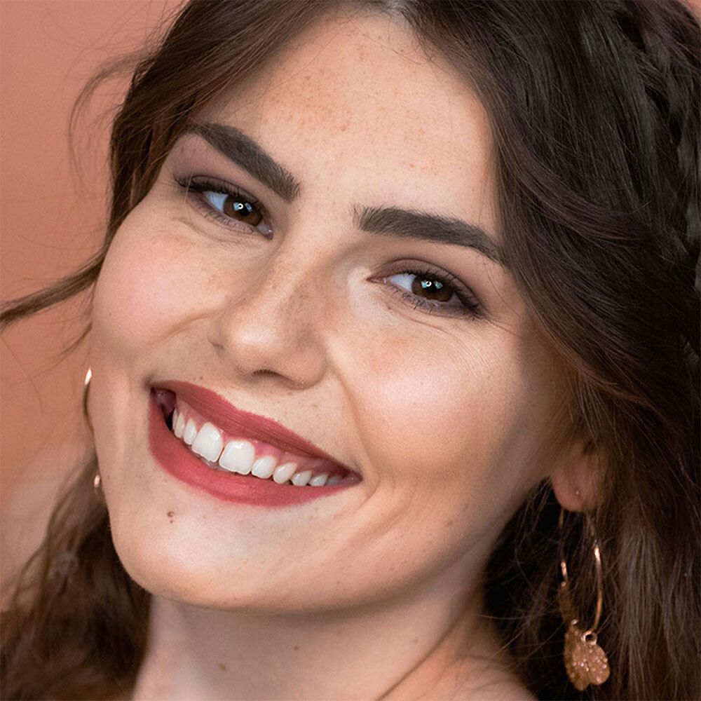 Portrait d'une femme aux sourcils dessinés. Elle porte du rouge à lèvres et des boucles d'oreilles. Peau avec des taches de rousseur.