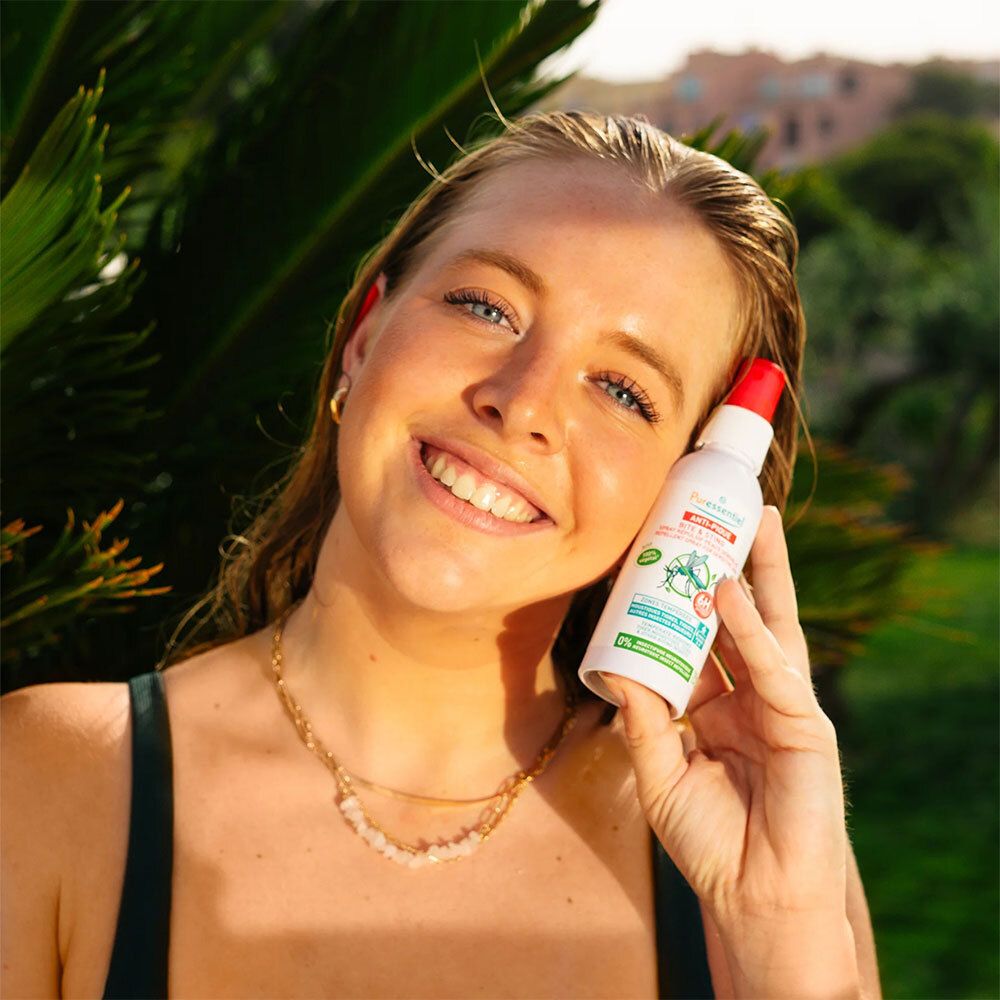 Jeune femme tenant un flacon spray blanc à bouchon rouge. Produit: Anti-Pique. Arrière-plan: plantes vertes.