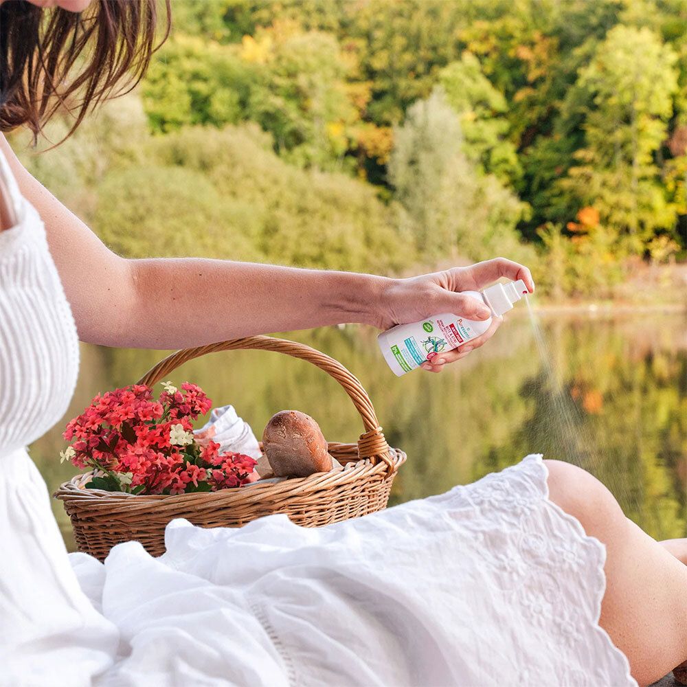 Femme vaporisant avec un flacon blanc à bouchon rouge. Produit: Anti-Pique. Arrière-plan: lac, panier avec fleurs.