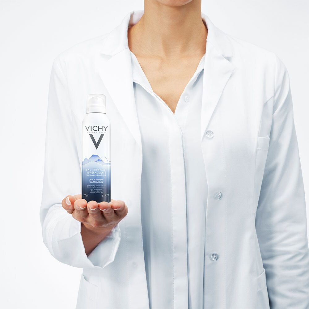 Personne tenant un spray d'eau thermale Vichy. Spray blanc avec design bleu. La personne porte une blouse blanche de laboratoire.