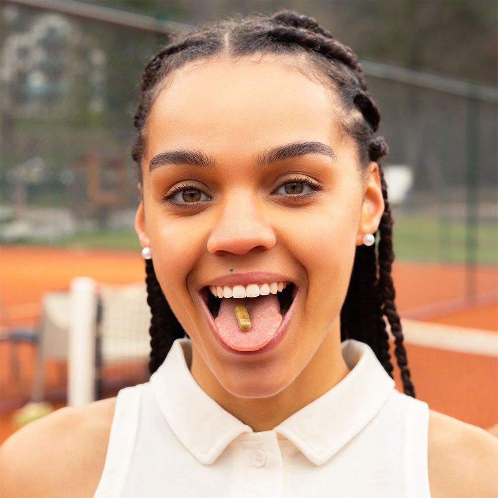 Femme avec une gélule dans la bouche. Articulations & Muscles SOS FLEX® Gélules. Fond : court de tennis.