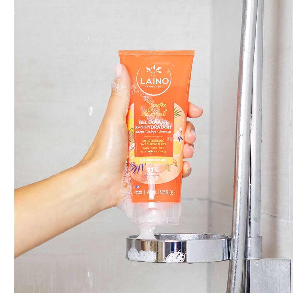 Main tenant un tube orange sous une pomme de douche. Nom du produit et logo visibles. Mousse autour du tube.