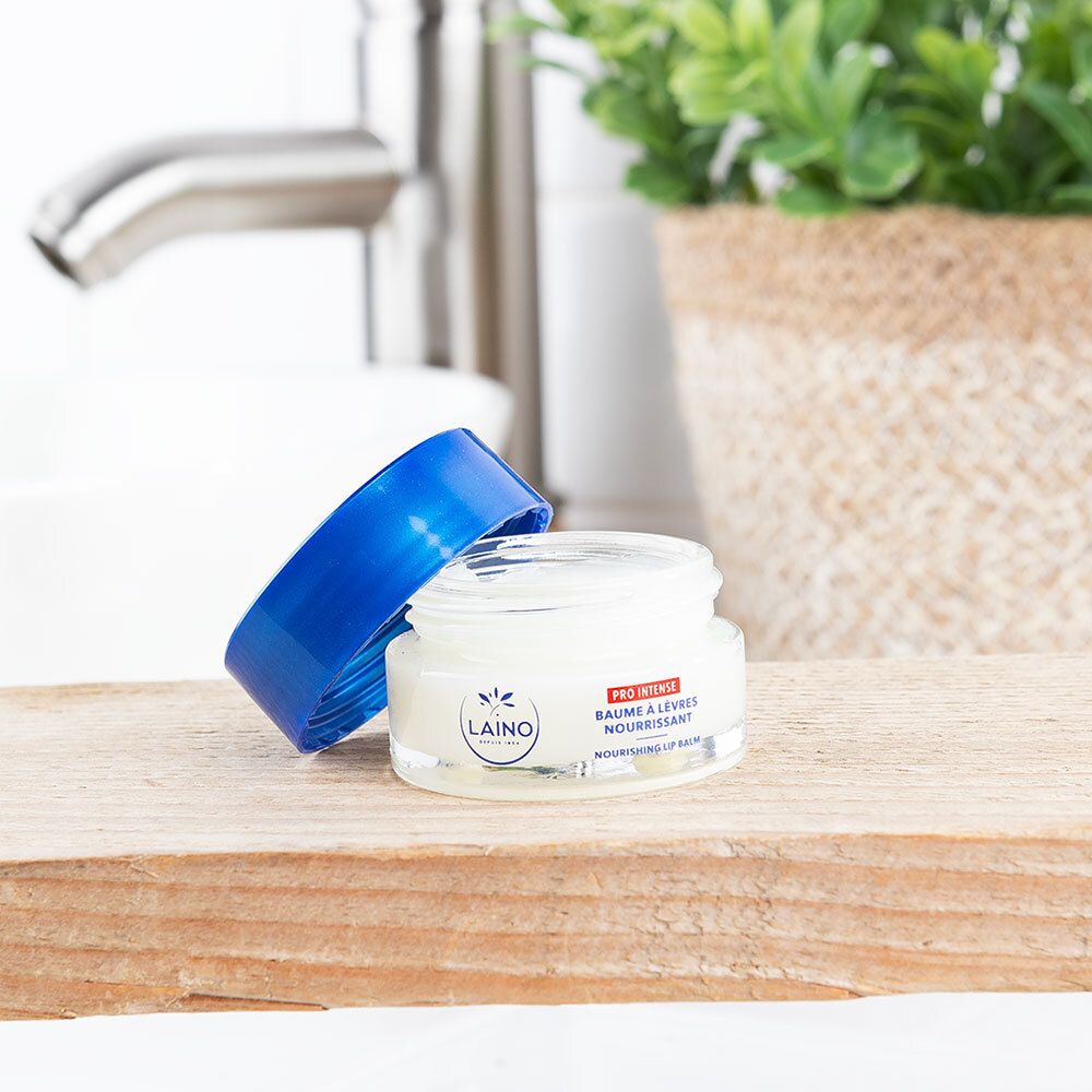 Pot de crème ouvert avec couvercle bleu. Inscription : LAINO. Posé sur du bois. En arrière-plan : lavabo et plante.