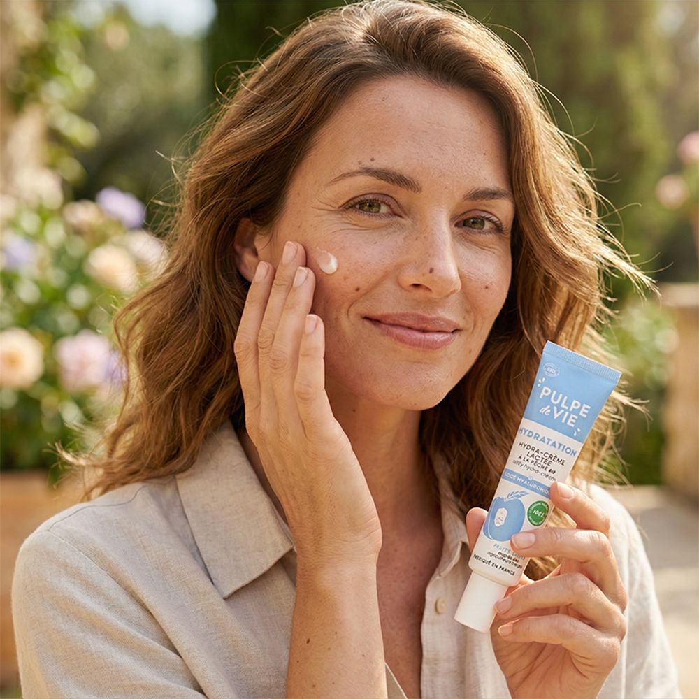 Femme tenant un tube avec le nom du produit. Elle applique de la crème sur son visage. Arrière-plan : jardin fleuri.