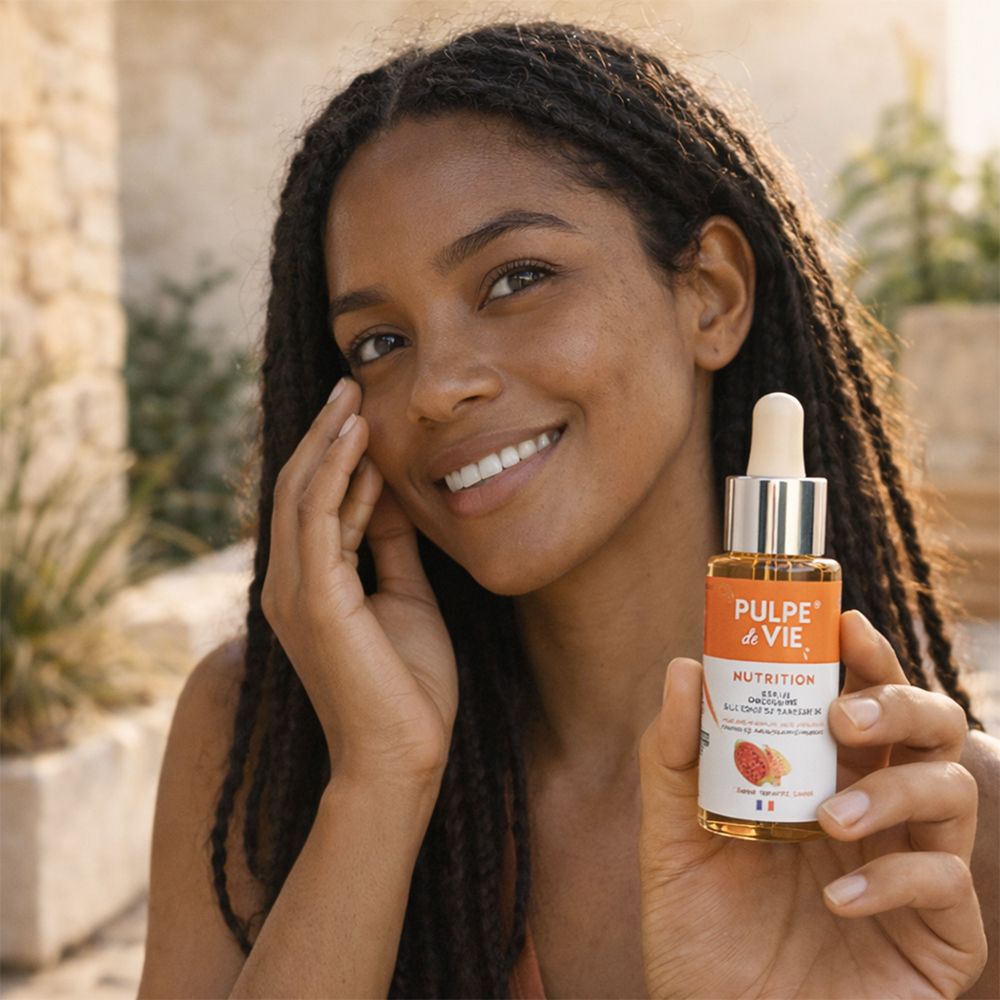 Femme tenant un flacon de sérum. Inscription: PULPE de VIE, NUTRITION. Sérum visage. La femme sourit. Fond: mur en pierre.