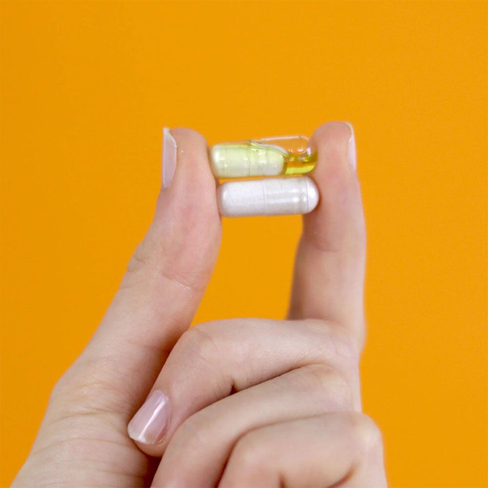Deux gélules dans une main. Gélule jaune et blanche. Fond orange.
