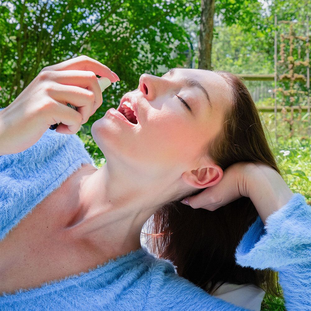 Femme vaporisant Puressentiel AROMA STRESS® Spray Buccal dans la bouche. Elle porte un peignoir bleu et a les yeux fermés.