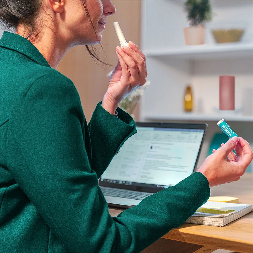 Une femme tient un inhalateur Puressentiel AROMA STRESS®. Un ordinateur portable et un autre inhalateur sur la table.
