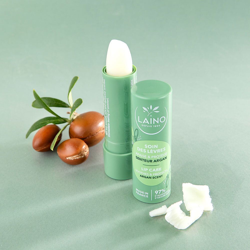 Stick de soin des lèvres ouvert, à côté d'argan et de feuilles. Texte: Laino, Soin des Lèvres, Senteur Argan, Lip Care. 