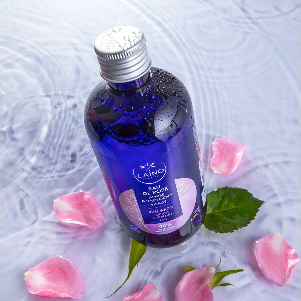 Flacon bleu avec bouchon argenté dans l'eau avec pétales de rose. Inscription: LAINO, Eau de Rose, apaise & rafraîchit le visage.